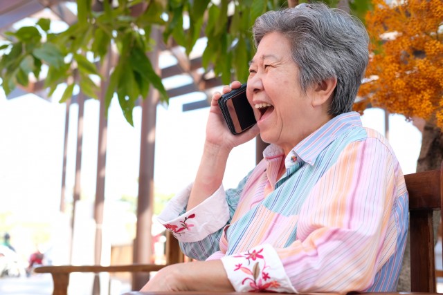 Äldre kvinna i trädgård som pratar i mobil. Kvinnan ler väldigt stort.
