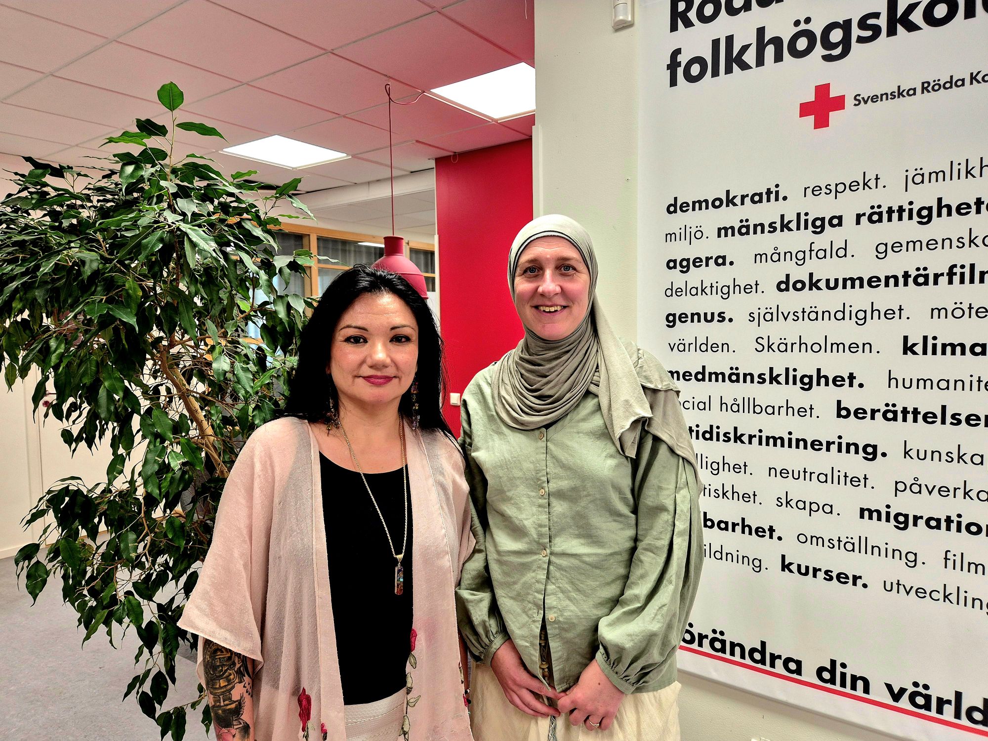 Kollegorna Grace och Anna på Röda Korsets folkhögskola framför en vägg med värdeord i Röds Korsets grafiska stil.