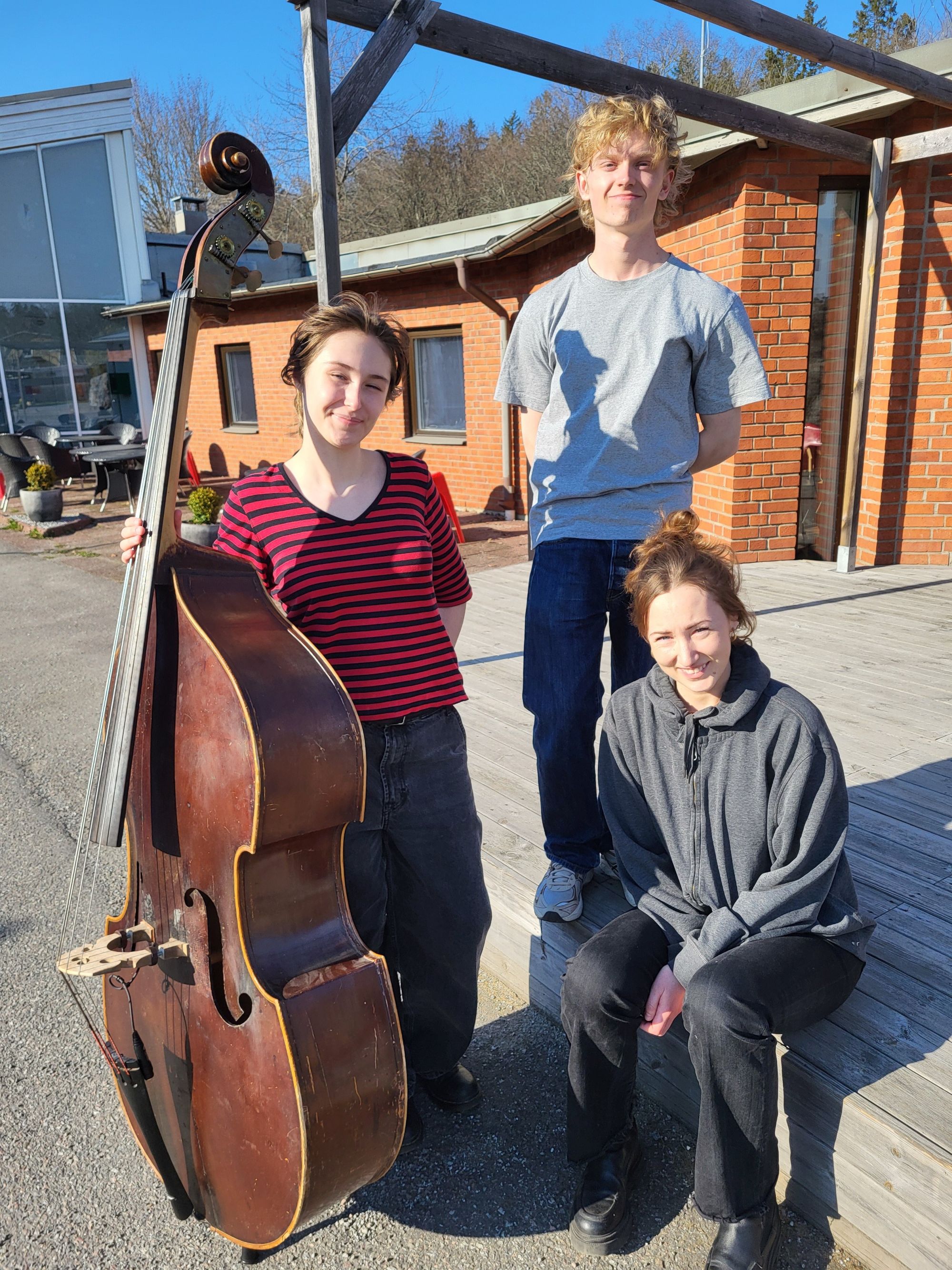 Tre deltagare på musiklinjen står i solskenet utanför Bromma folkhögskola med en kontrabas.
