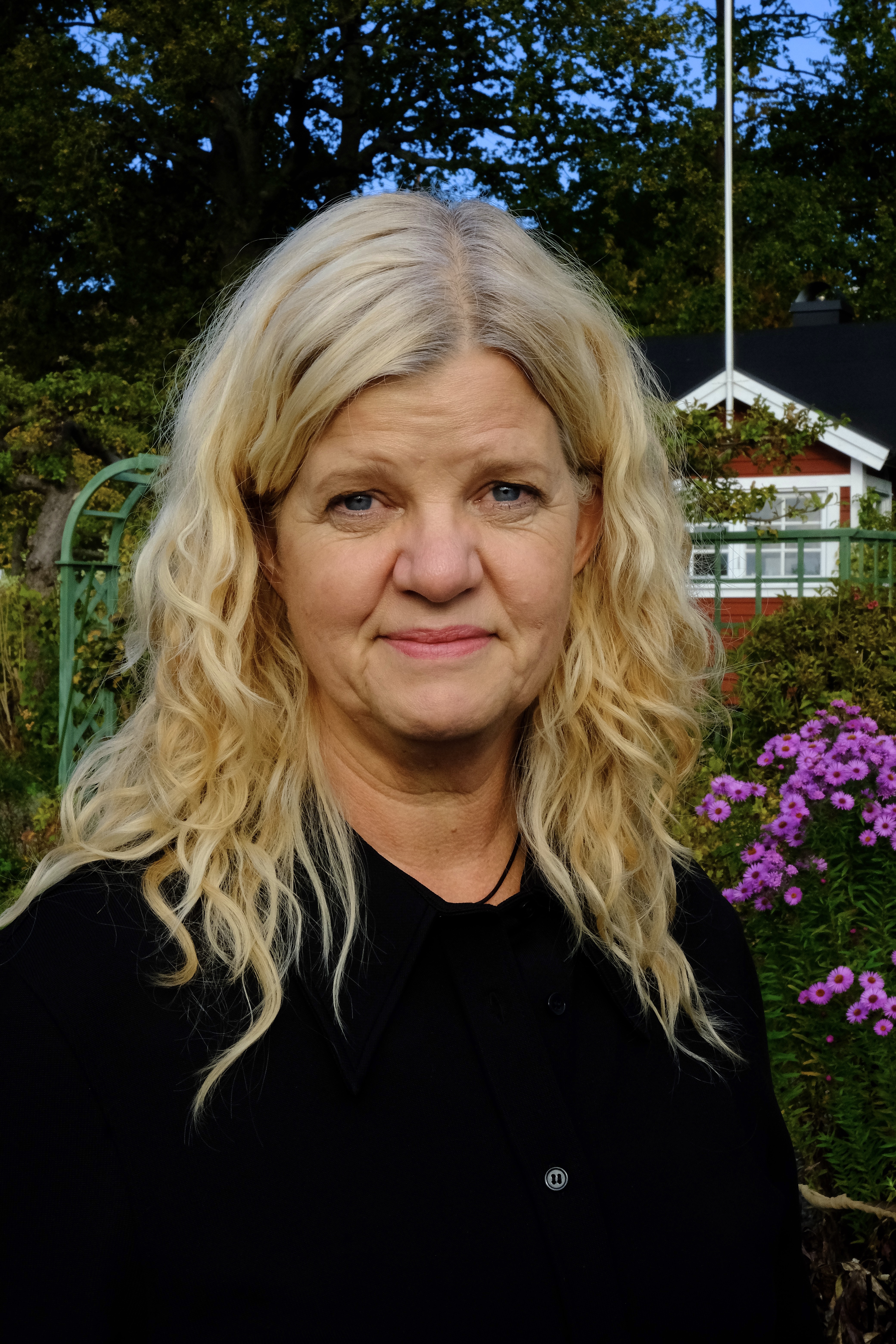 Ulrica står med en koloniträdgård med lila blommor och en röd stuga med vita knutar i bakgrunden.