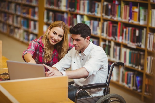 Två personer i ett bibliotek som studerar framför en dator. En av dem sitter i rullstol.