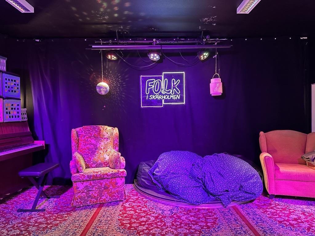 Två fåtöljer står på en scen i ett rum med lila belysning. På väggen hänger en skylt där det står "Folk i Skärholmen".