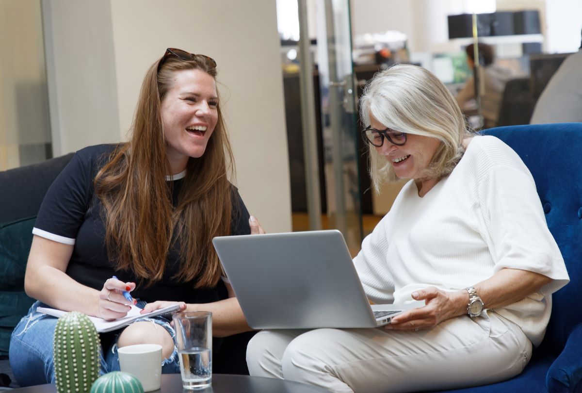 Två kvinnor skrattar när de diskuterar arbete, sittande i soffa i på kontoret med laptop i knät.