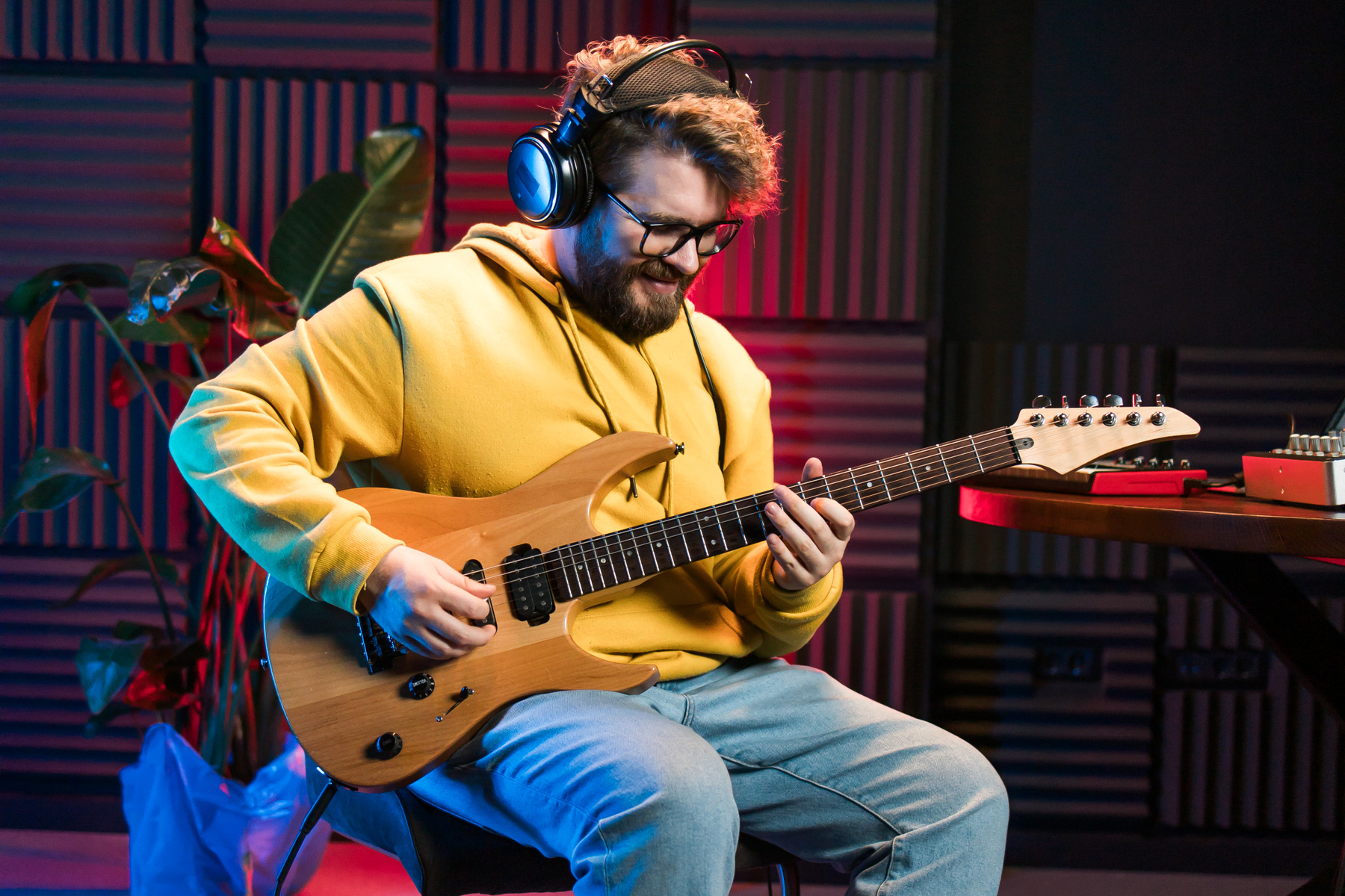 Man sitter i studio och spelar gitarr. Han har hörlurar på sig, rött hår och skägg, och en gul hoodie.