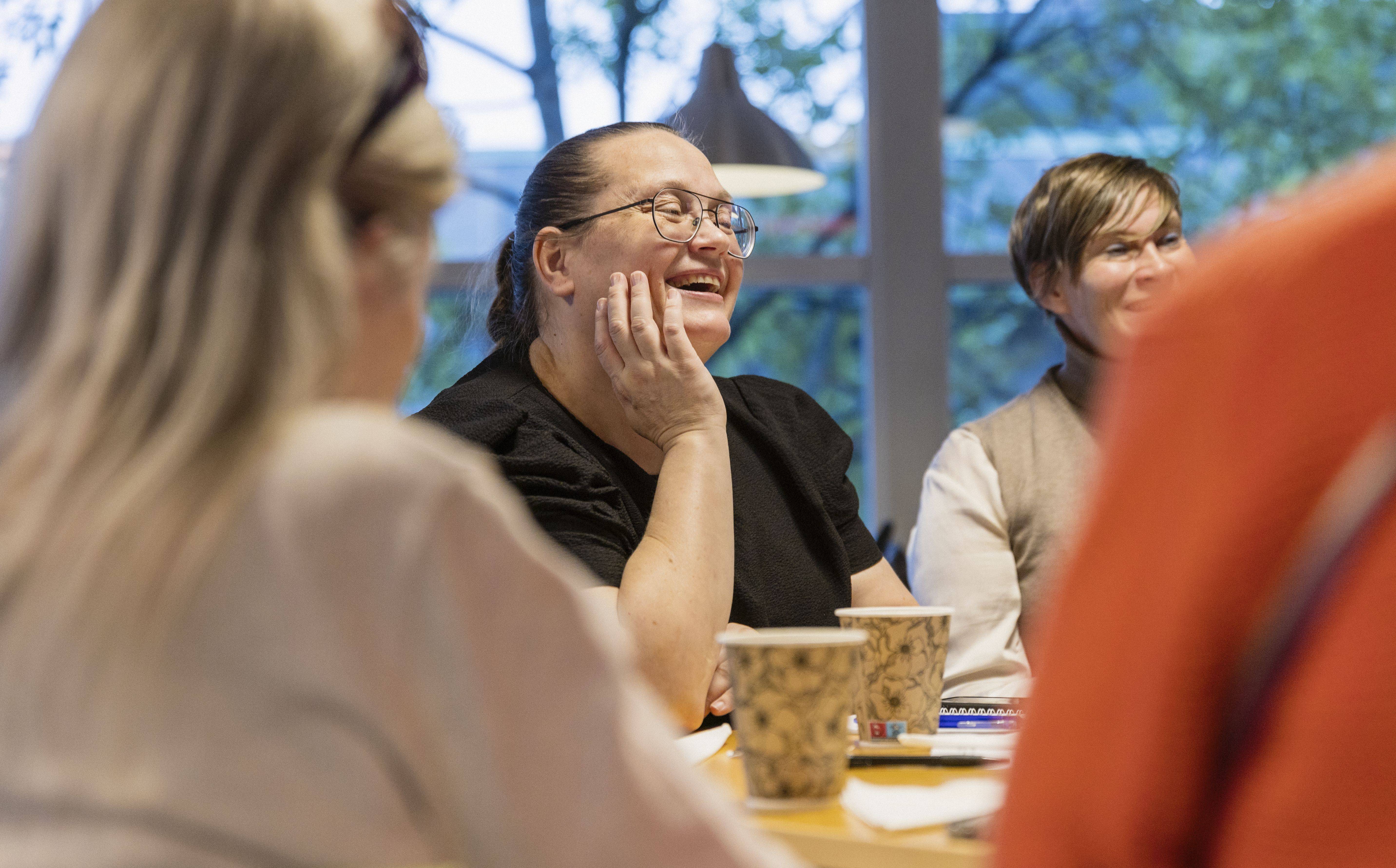 Fyra personer runt ett bord. En person syns framifrån och ler. På bordet står två pappersmuggar bland papper och pennor.