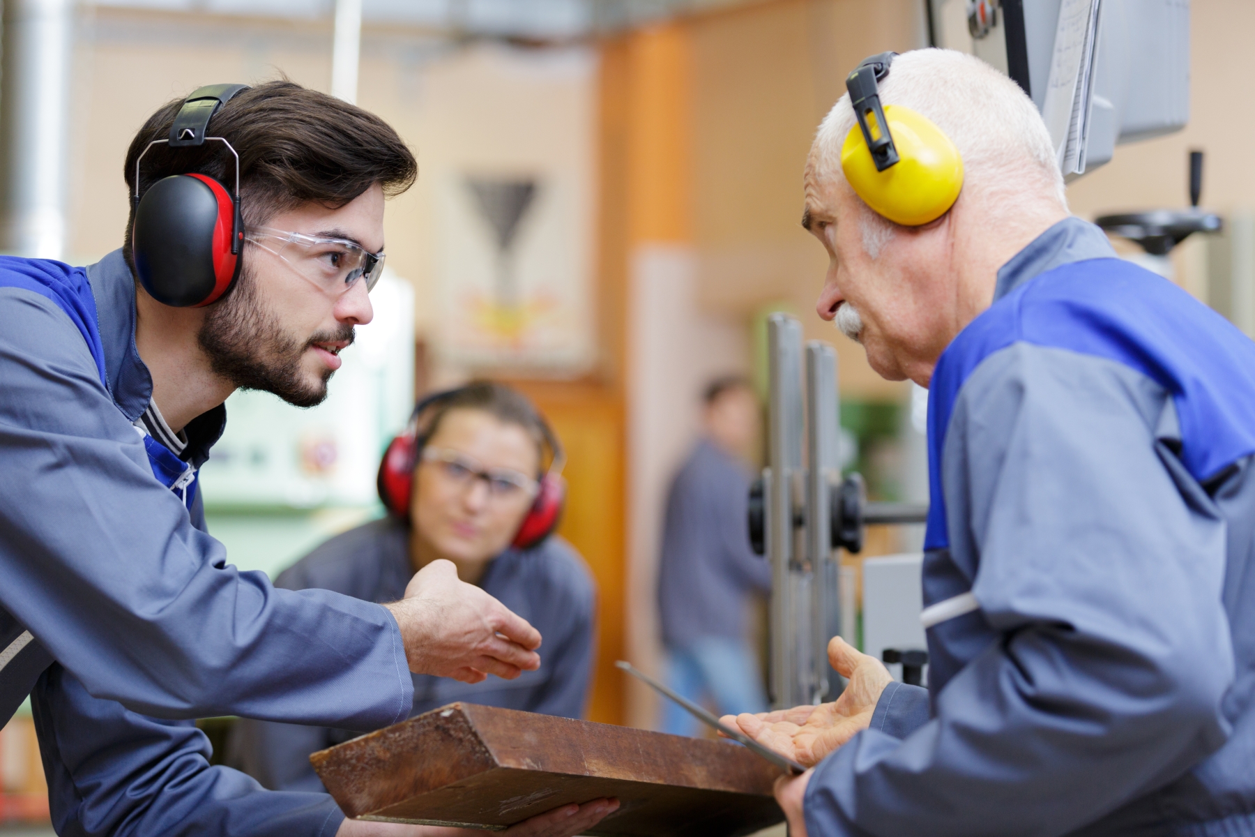 En yngre och en äldre man står och diskuterar iklädda hörselskydd och skyddskläder. 