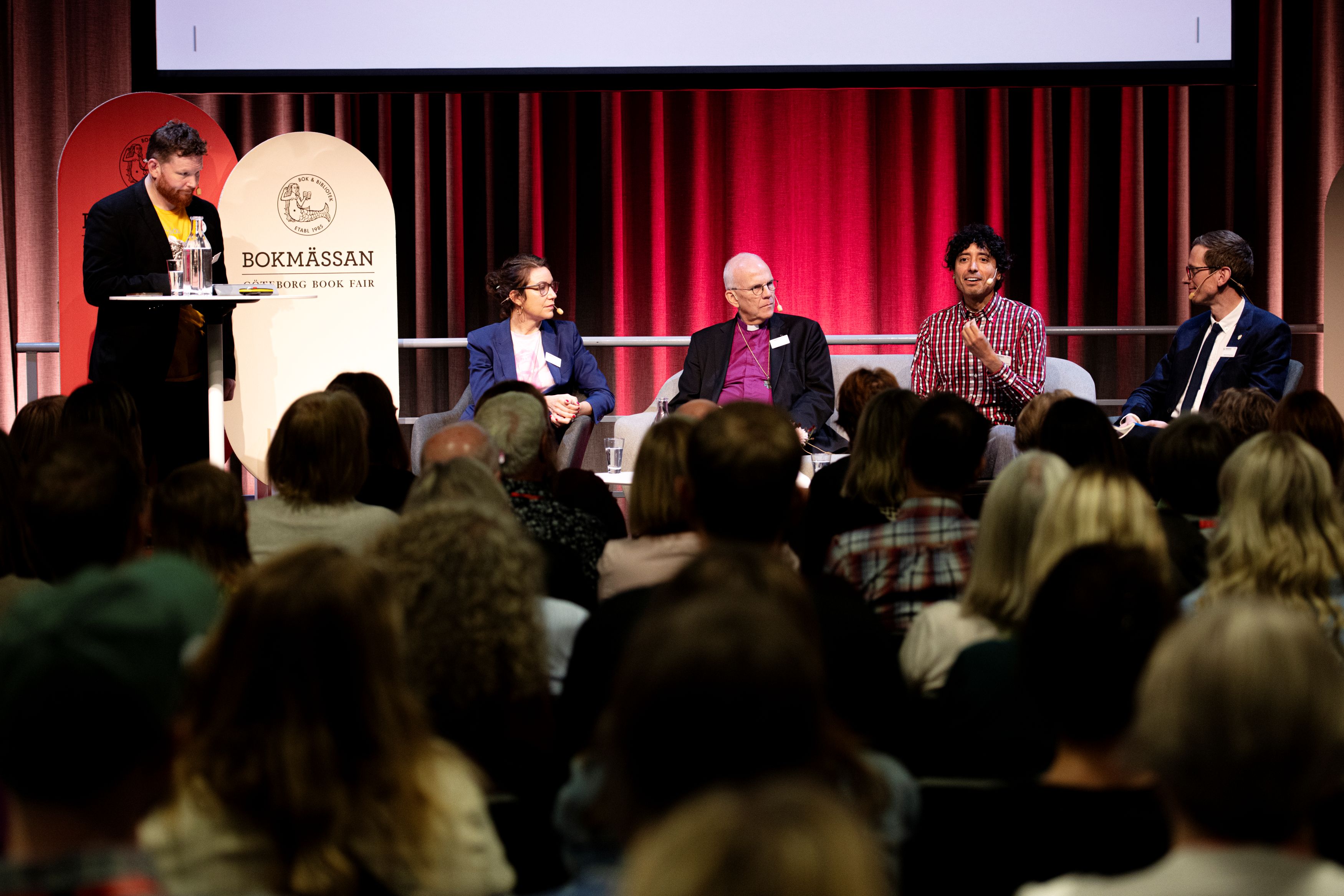Seminarium på Bokmässan. Moderator och panel på scenen med rött draperi i bakgrunden. Foto: Magnus Liam Karlsson