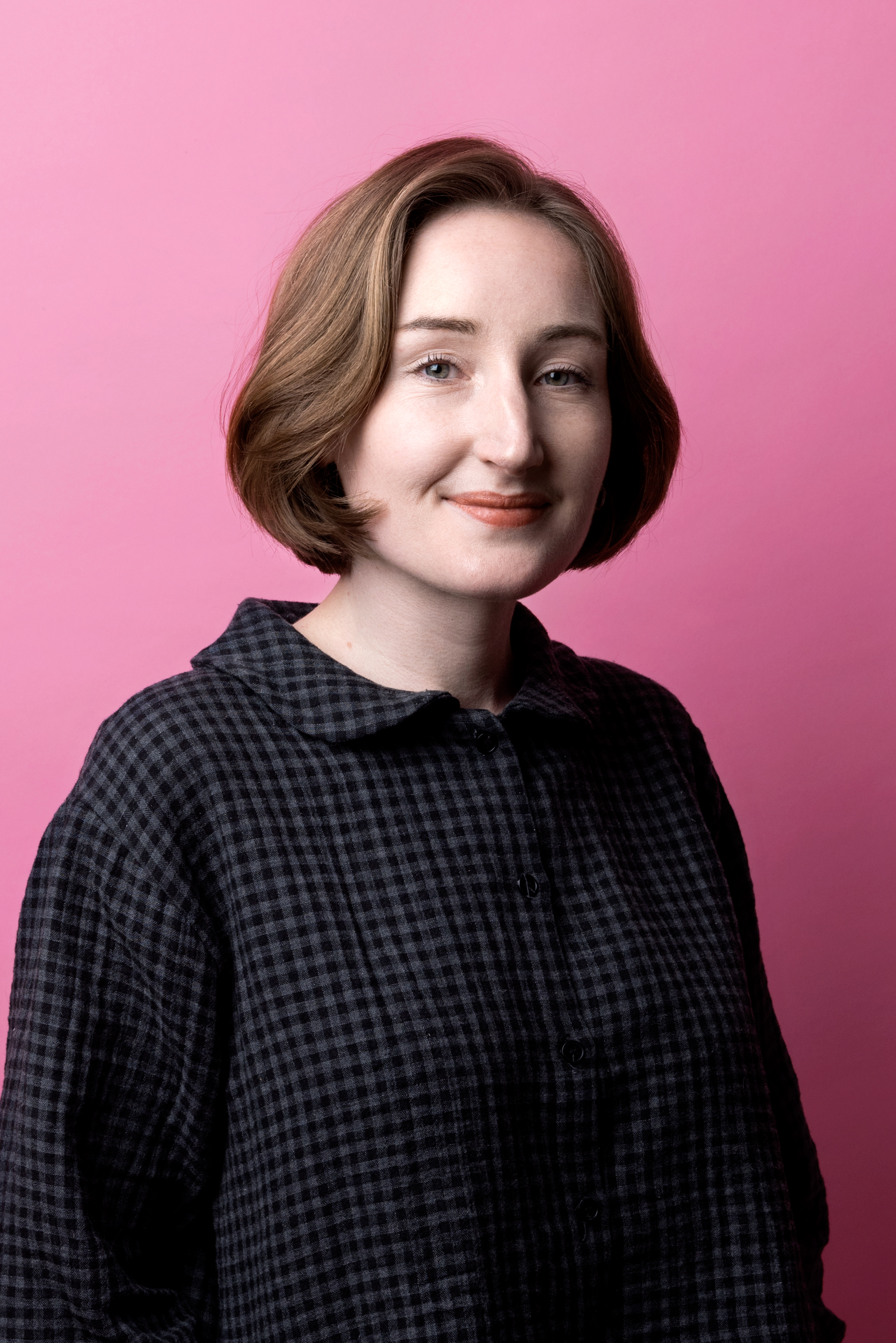 Porträttbild Ella Coogan. Ella har brunt hår i page och en mörk rutig blus. Hon tittar in i kameran med ett litet leende.